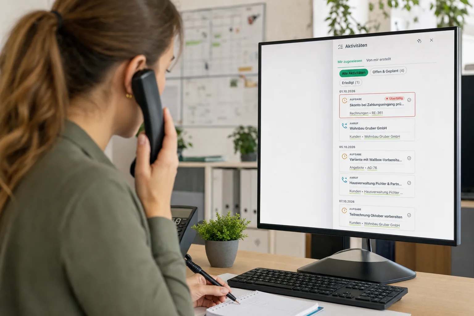 Mitarbeiterin im Büro telefoniert und notiert mit, am Monitor die Aufgabenliste mit einer überfälligen Aufgabe an einer Rechnung